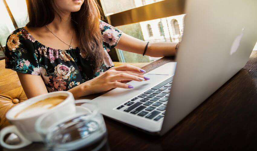 Eine Frau sitzt vor einem Laptop. Neben dem Laptop eine Tasse Caffee und ein Glas Wasser