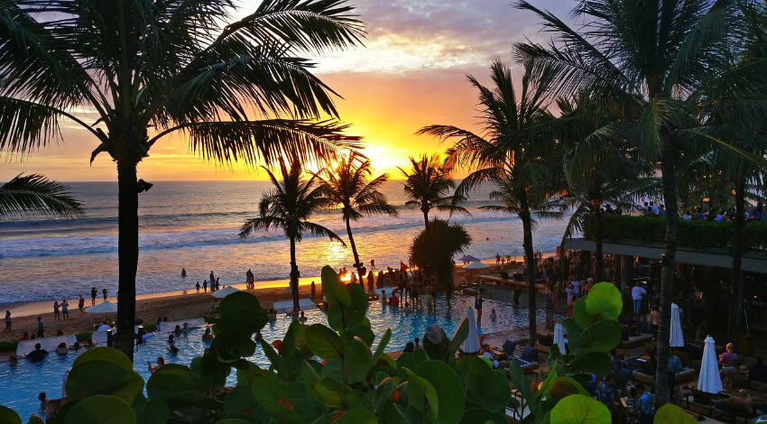 Strand auf Bali mit Palmen und Restaurant bei Sonnenuntergang.