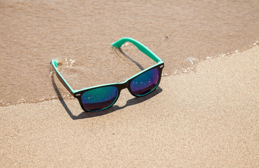 Eine Sonnenbrille mit türkisen Bügeln lieg an einem Sandstrand.