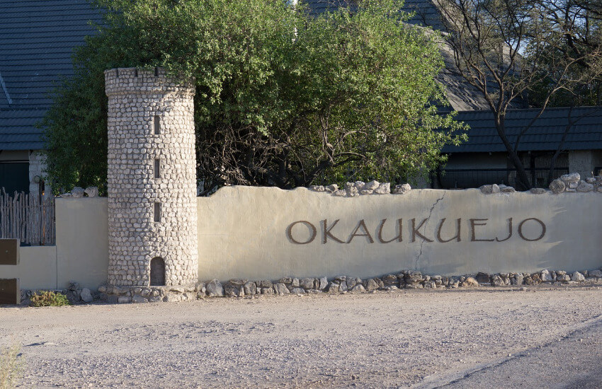 Ein kleiner Turm aus Steinen, rechts davon eine Mauer mit der Aufschrift Okaukuejo.