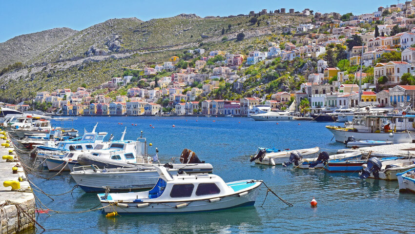 Boote ankern am Hafen Symi und im Hintergrund ein felsiger Hügel mit Häusern.