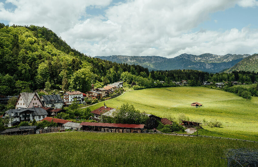 Idyllisches Bergdorf mit grünen Wiesen, Waldhängen und Alpenpanorama unter Wolken.