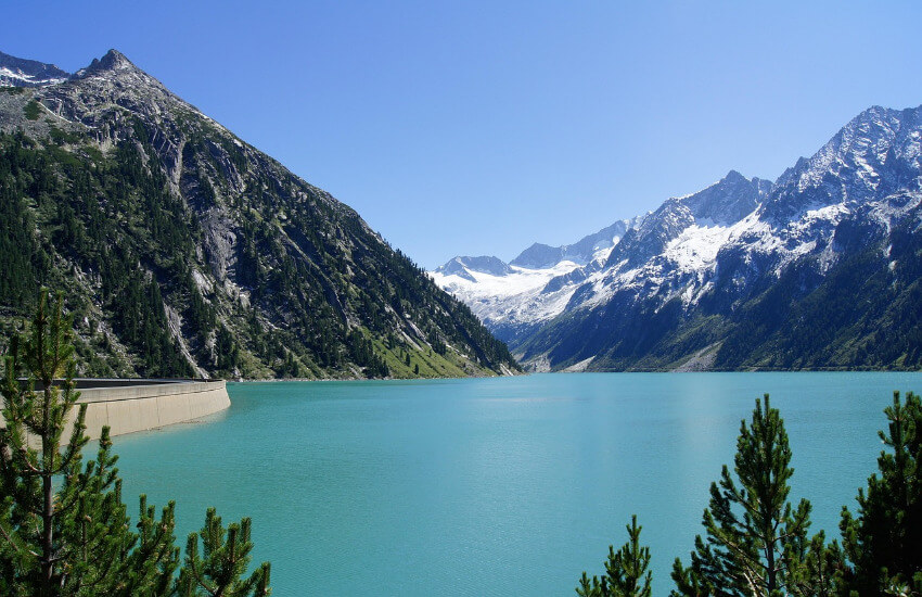 Kleine Hängebrücke aus Holz und Stahlseilen über einem Gebirgsbach mit spektakulärem Blick auf den türkisfarbenen Schlegeis-Stausee und die Zillertaler Alpen.