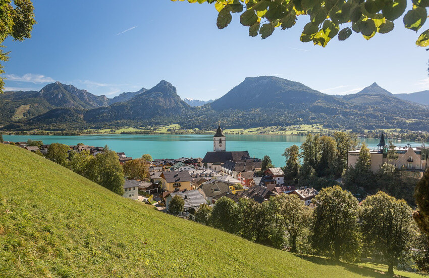 Ein türkiser See, im Vordergrund ein Dorf mit Kirchturm und im Hintergrund Berge.