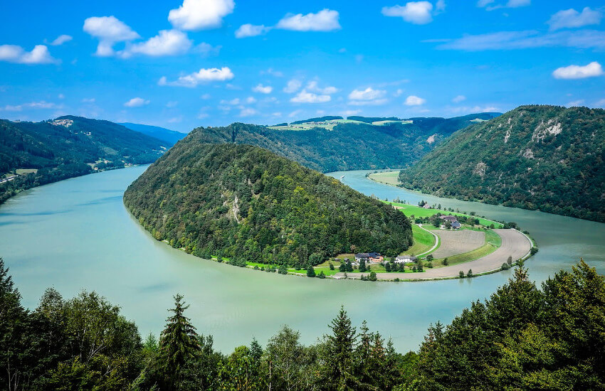 Luftaufnahme der Schlögener Schlinge, einer markanten 180-Grad-Kurve der Donau in Oberösterreich. Der Fluss windet sich in einer tiefen S-Kurve durch bewaldete, grüne Hügel. Am linken Flussufer ist die kleine Ortschaft Schlögen mit einem Schiffsanleger zu sehen.